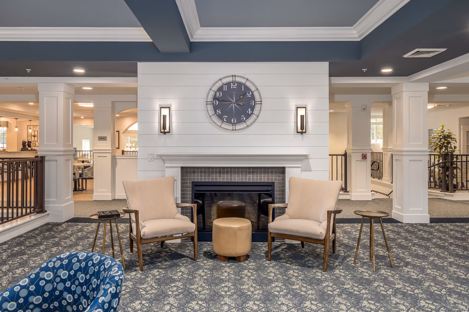 A cozy senior living community lounge area featuring two beige armchairs with wooden arms, a small round tan ottoman between them, and two small round side tables with gold legs. Behind the seating is a white shiplap wall with a modern fireplace and a large round clock above it. The ceiling is painted blue with white crown molding, and the floor is covered with a patterned carpet. The space is well-lit with wall sconces and recessed lighting, and there are white columns and railings in the background.