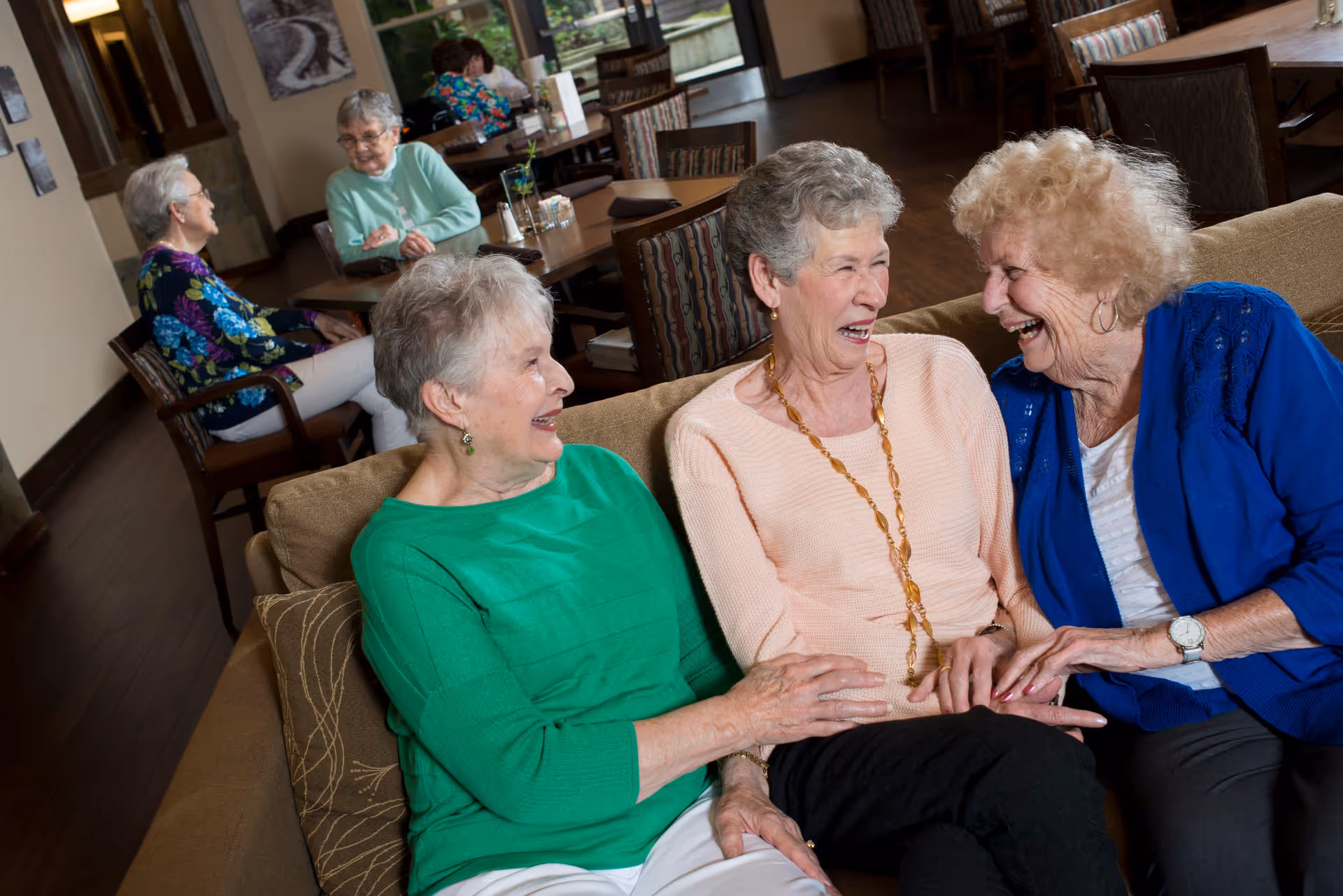 Three elderly women sitting on a couch in a common area, laughing and enjoying each other's company. In the background, two other elderly women are seated at a table engaged in conversation. The room has wooden floors, tables, chairs, and large windows letting in natural light.