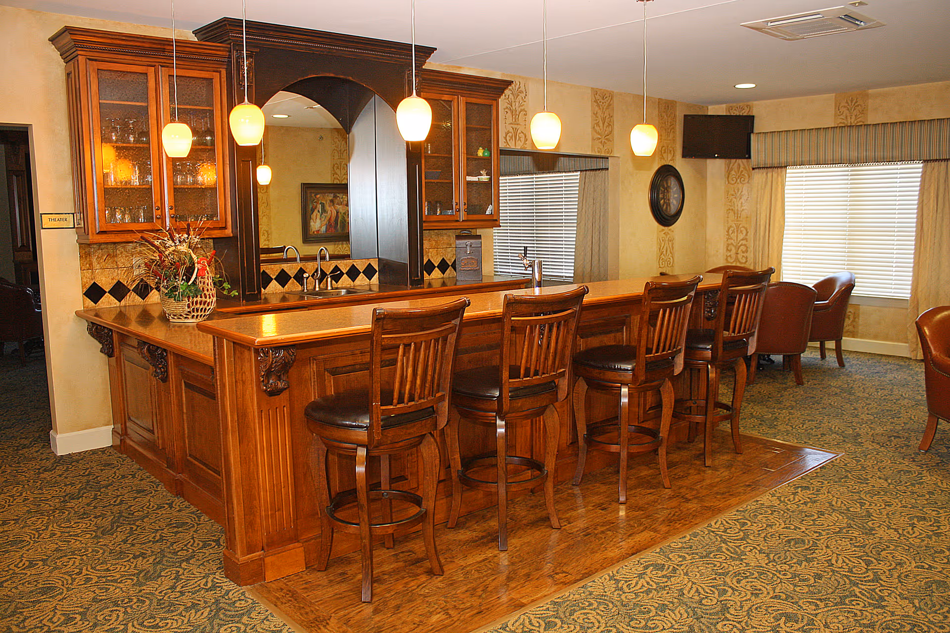 Interior view of a senior living facility lounge area featuring a wooden bar counter with five high wooden chairs. The bar has a built-in sink and glass cabinets above it. Pendant lights hang from the ceiling above the bar. The room has patterned carpet, beige walls with decorative wallpaper, a wall clock, a mounted TV, and windows with blinds and valances. There are also a few leather armchairs in the background.