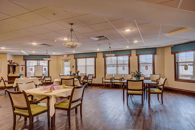 Bright communal dining room with multiple tables and chairs, wood flooring, large windows, and hanging light fixtures.