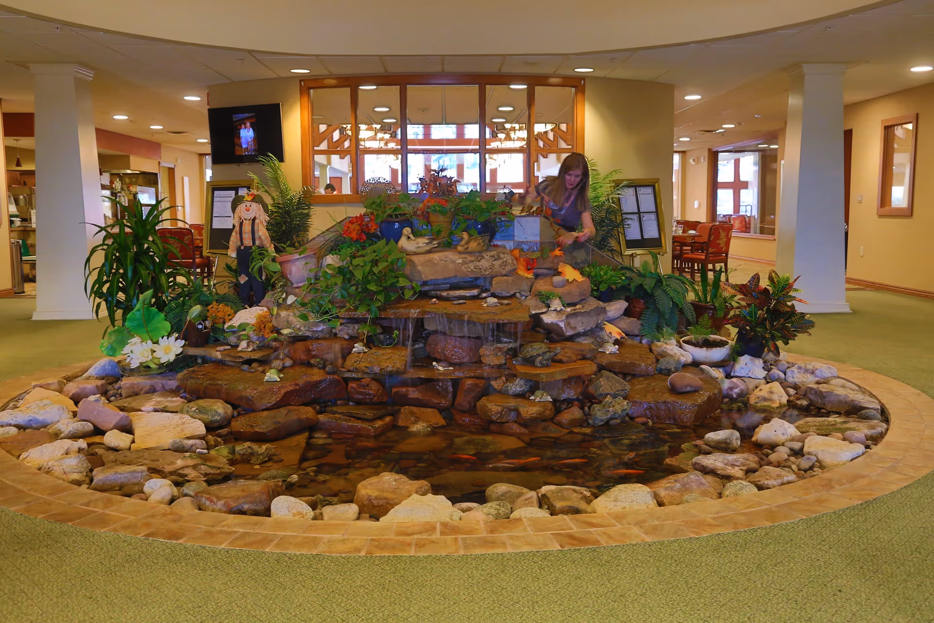 Indoor water feature with rocks, plants, and a small pond in the center of a room. A woman is tending to the plants on the water feature. The room has green carpet, columns, and tables with chairs in the background.