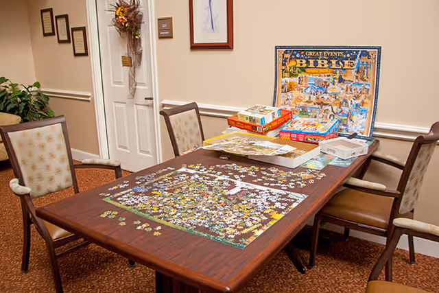 A wooden table in a room with four chairs around it. On the table, there is a partially completed jigsaw puzzle with scattered puzzle pieces, several puzzle boxes, and a completed puzzle image titled 'Great Events Bible'. The room has beige walls, a carpeted floor, framed pictures on the walls, and a door decorated with a wreath.