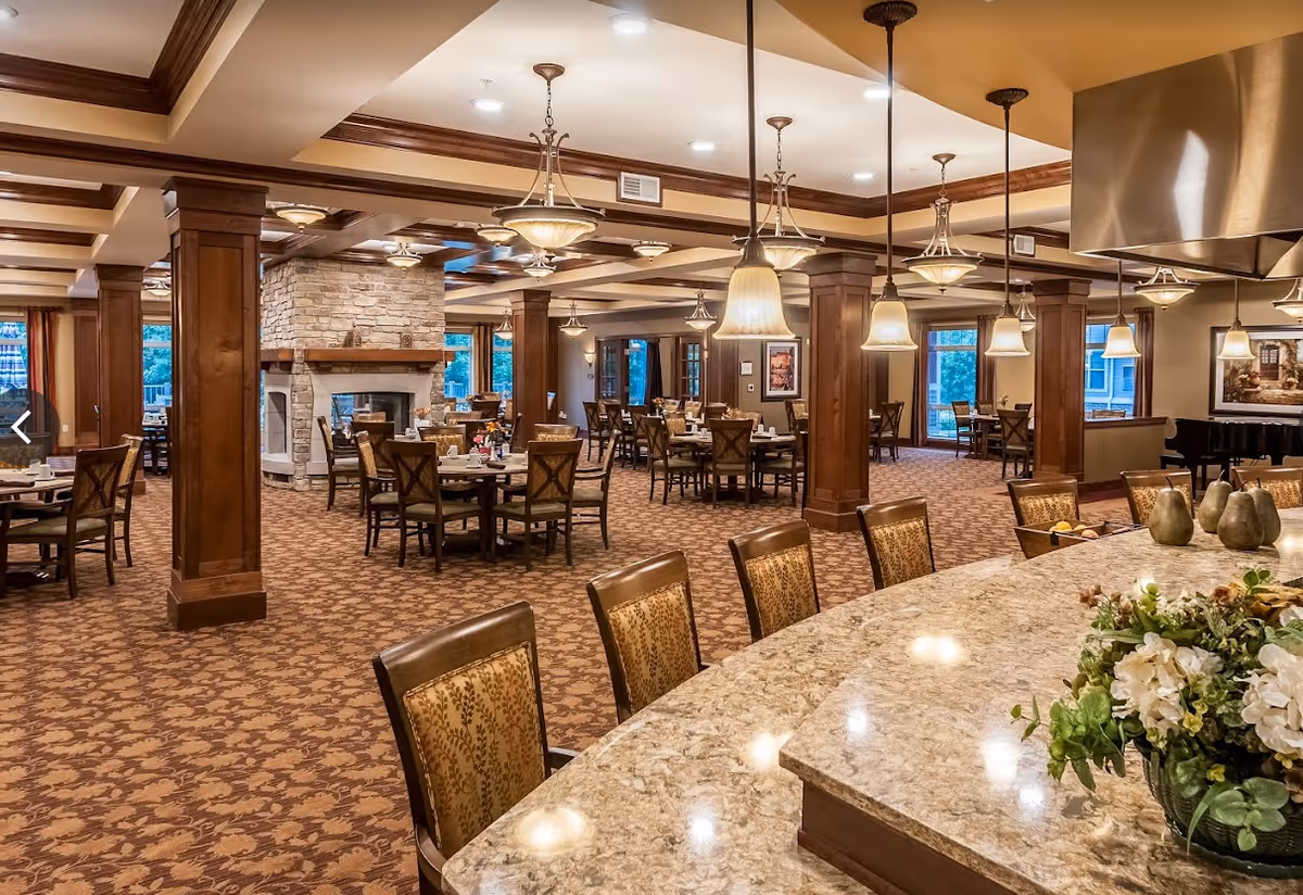 Spacious dining area with multiple wooden tables and chairs arranged neatly on a patterned carpet. The room features a large stone fireplace in the center, decorative ceiling lights, and large windows letting in natural light. In the foreground, there is a granite countertop with floral arrangements and decorative vases.