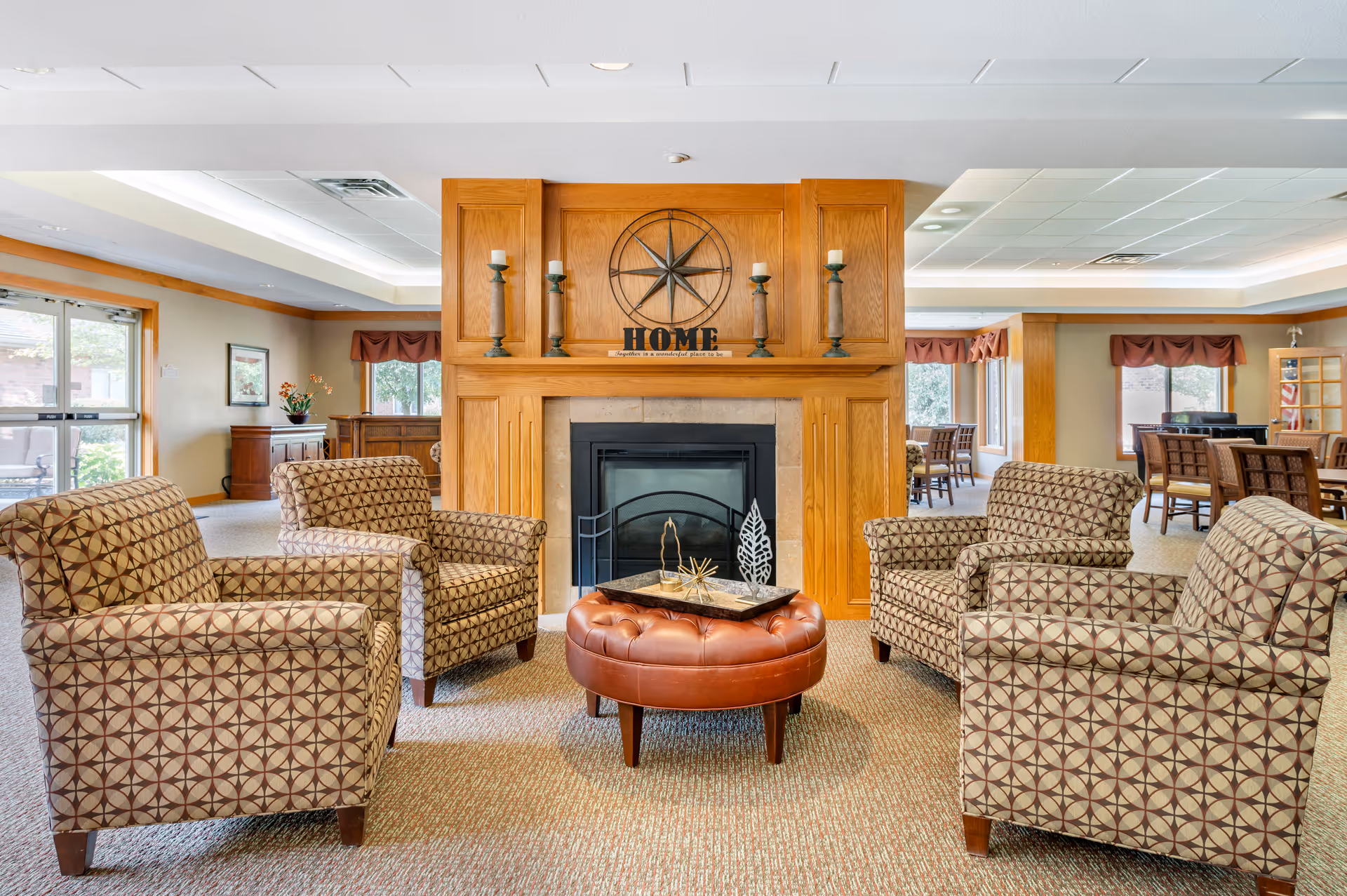 A cozy common area in a senior living facility featuring four patterned armchairs arranged around a round brown leather ottoman with decorative items on top. Behind the seating area is a wooden fireplace mantel with candles and a metal compass wall decoration above the word 'HOME'. Large windows and additional seating areas are visible in the background.