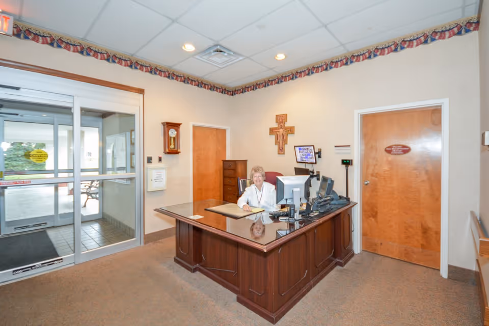 Reception area of a senior living facility with a receptionist seated behind a wooden desk near automatic glass entrance doors.
