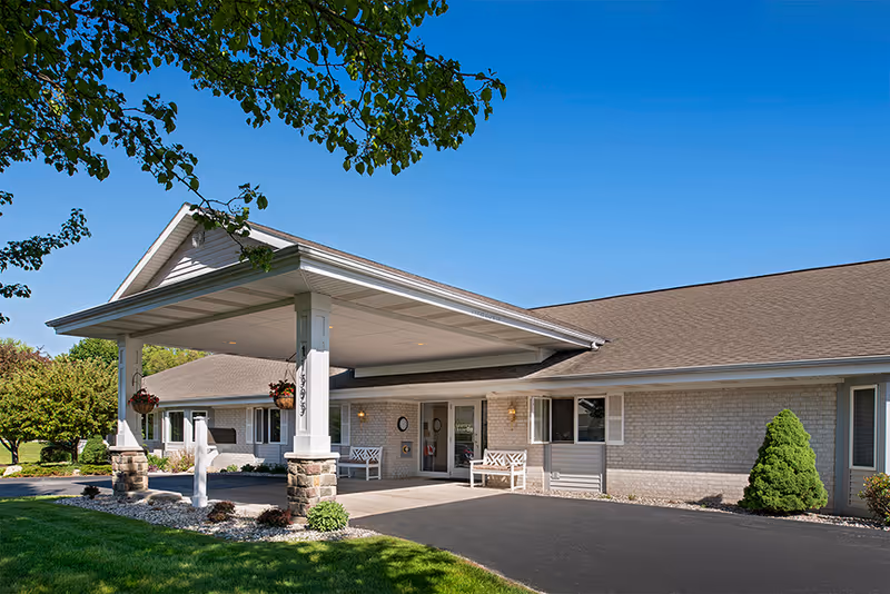 Exterior view of American House Spring Lake facility showing a single-story building with a covered entrance, benches, hanging flower pots, and a well-maintained lawn under a clear blue sky.