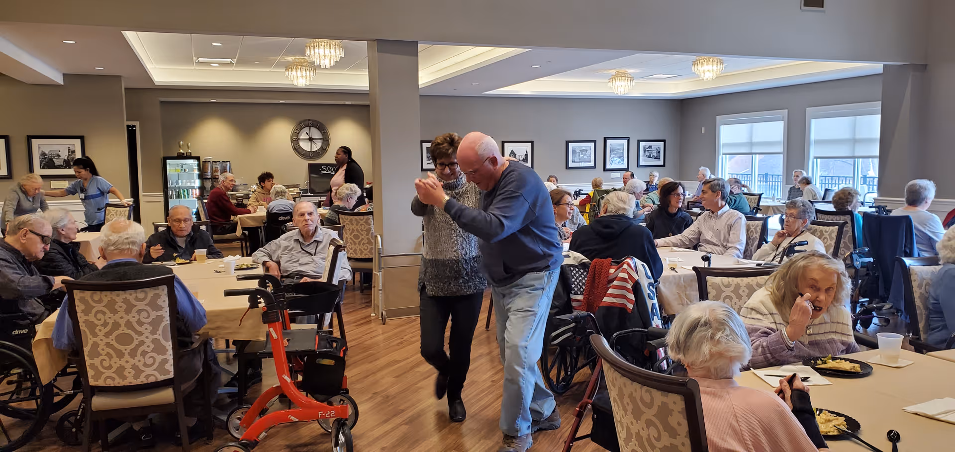 A lively dining room in a senior living facility where elderly residents are seated at tables eating and socializing. In the center, an elderly couple is dancing together. The room has large windows with natural light, framed pictures on the walls, and a wooden floor. Staff members are assisting residents in the background.