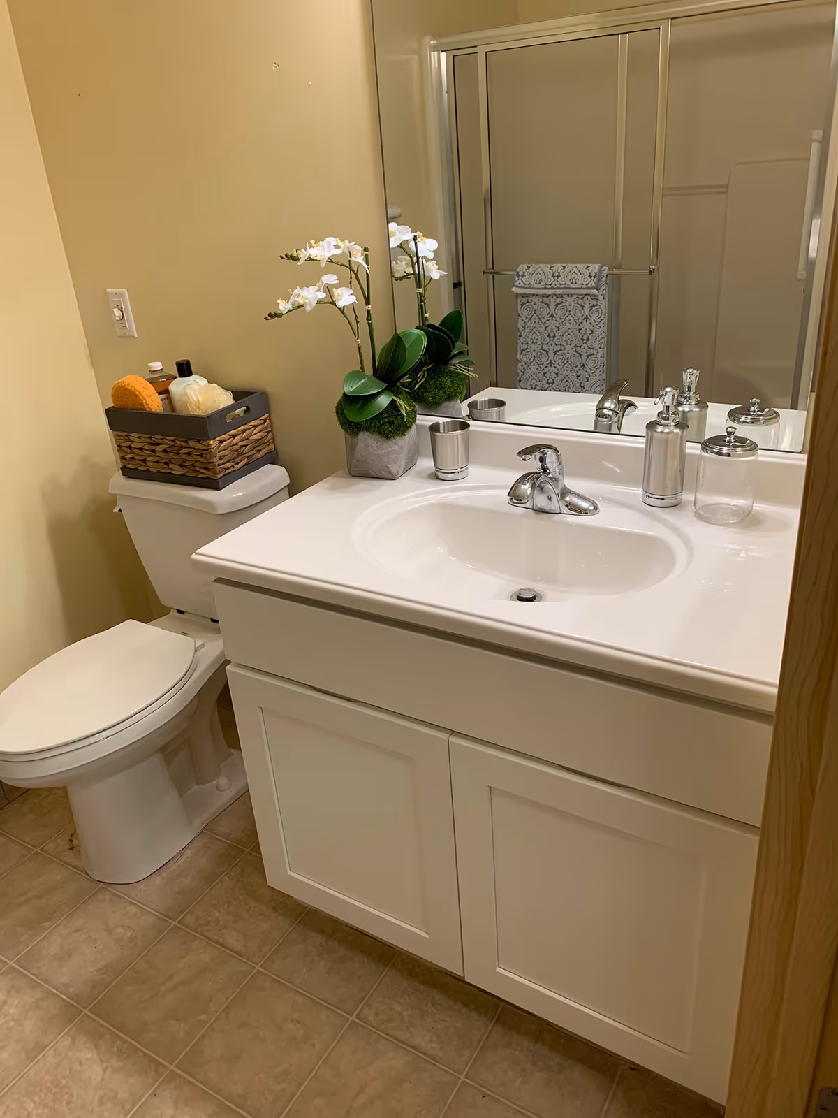 A clean bathroom with a white sink and cabinet, a large mirror above the sink, a toilet with a basket containing toiletries on top, a decorative white orchid plant on the sink counter, and a glass shower enclosure with a patterned towel hanging on the door.