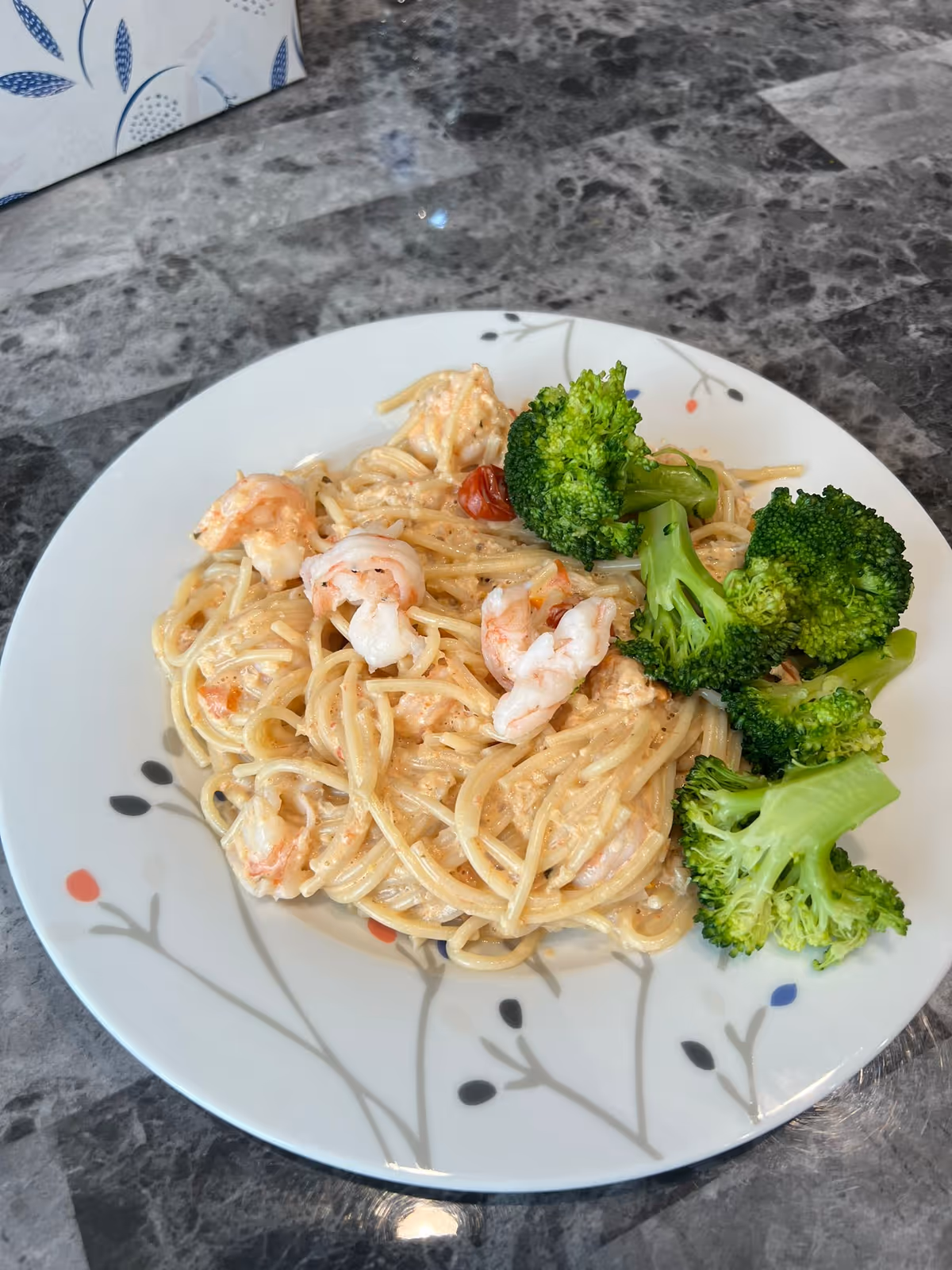 Plate of shrimp pasta with broccoli on a patterned white plate on a gray marble table.