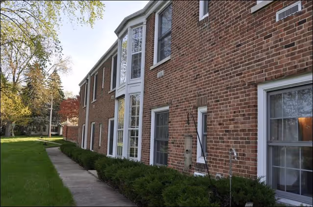 Exterior view of a two-story brick building with multiple windows, a concrete walkway, and green shrubs along the side. Trees and grass are visible in the surrounding area under a clear sky.