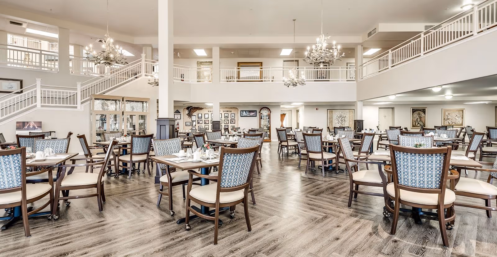 Spacious dining room with multiple wooden tables and chairs featuring patterned upholstery. The room has a high ceiling with two chandeliers, a staircase leading to an upper level with white railings, and framed artwork on the walls. The floor is a light wood pattern, and the tables are set with white napkins and small flower arrangements.