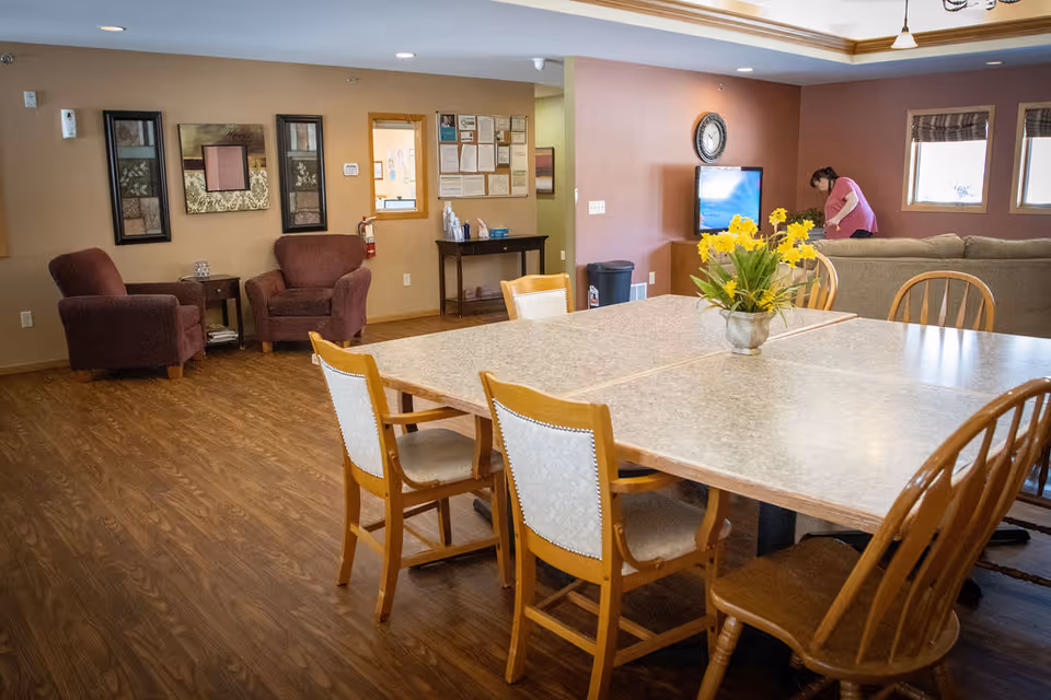 A communal room with a large table surrounded by wooden chairs, a vase with yellow flowers on the table, two armchairs against a wall with framed artwork, a television on a stand, and a person in a pink shirt near a window in the background.