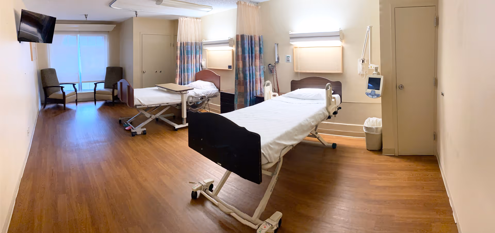 Large senior-care patient room with two adjustable hospital beds, a curtain divider, seating area by the window, wall-mounted TV, and wood-look flooring.