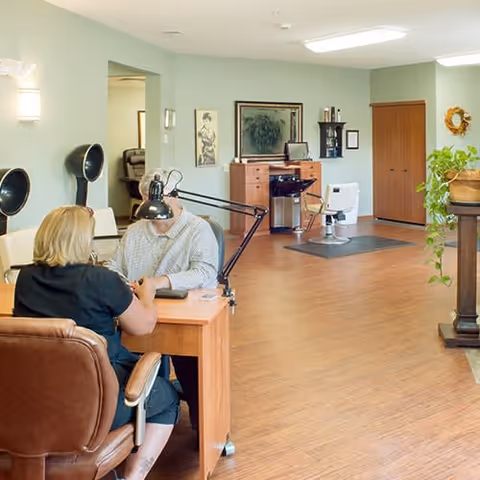 A senior living facility interior showing a woman providing a manicure to a man seated at a wooden table. The room has light green walls, wooden flooring, and salon equipment including hair dryers and a styling chair. There are framed pictures and plants decorating the space.