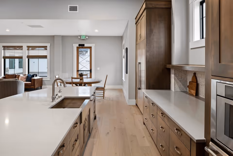 Modern kitchen area with a large white island countertop featuring a built-in sink and faucet. Wooden cabinets line the right side with a built-in oven and refrigerator. In the background, there is a small dining table with chairs and a door with glass panels. A living room area with sofas and windows is visible to the left.