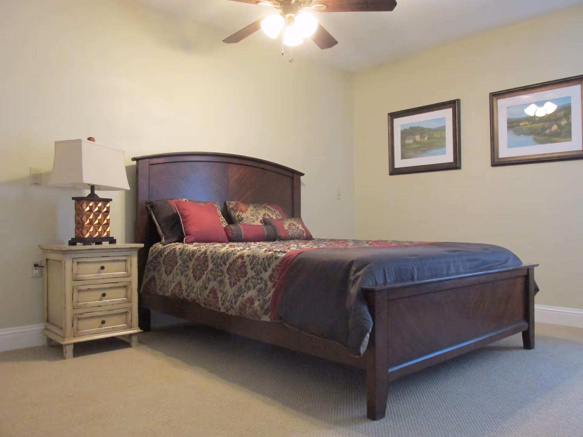 A bedroom with a wooden bed frame and a bed made with patterned bedding in shades of red, gray, and beige. There is a nightstand with a lamp on the left side of the bed. Two framed landscape paintings hang on the wall above the bed. A ceiling fan with lights is visible on the ceiling.