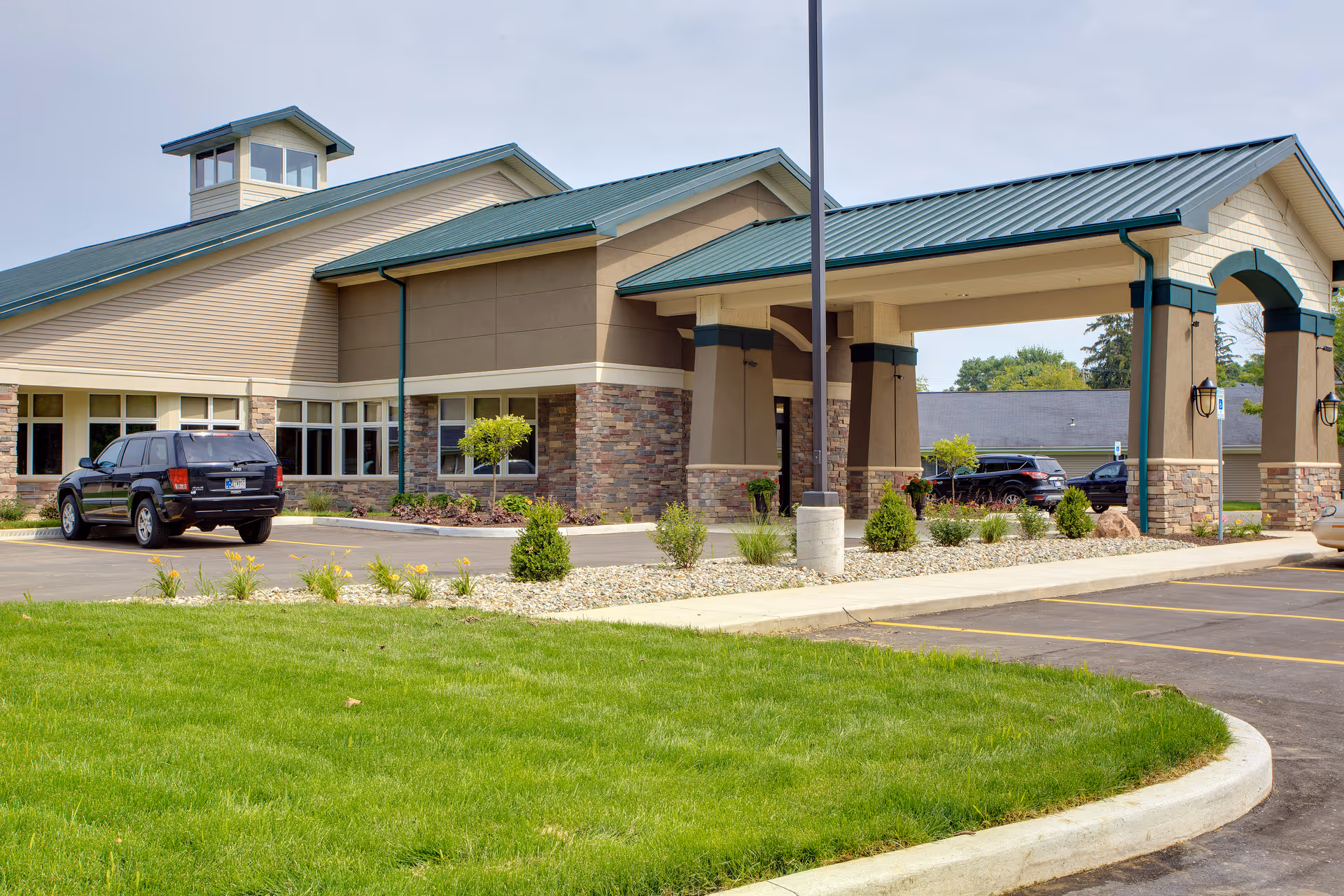 Exterior view of Miller's Merry Manor facility showing the entrance with a covered drop-off area, stone and beige siding, green metal roof, parked cars, and landscaped greenery with grass and small bushes.