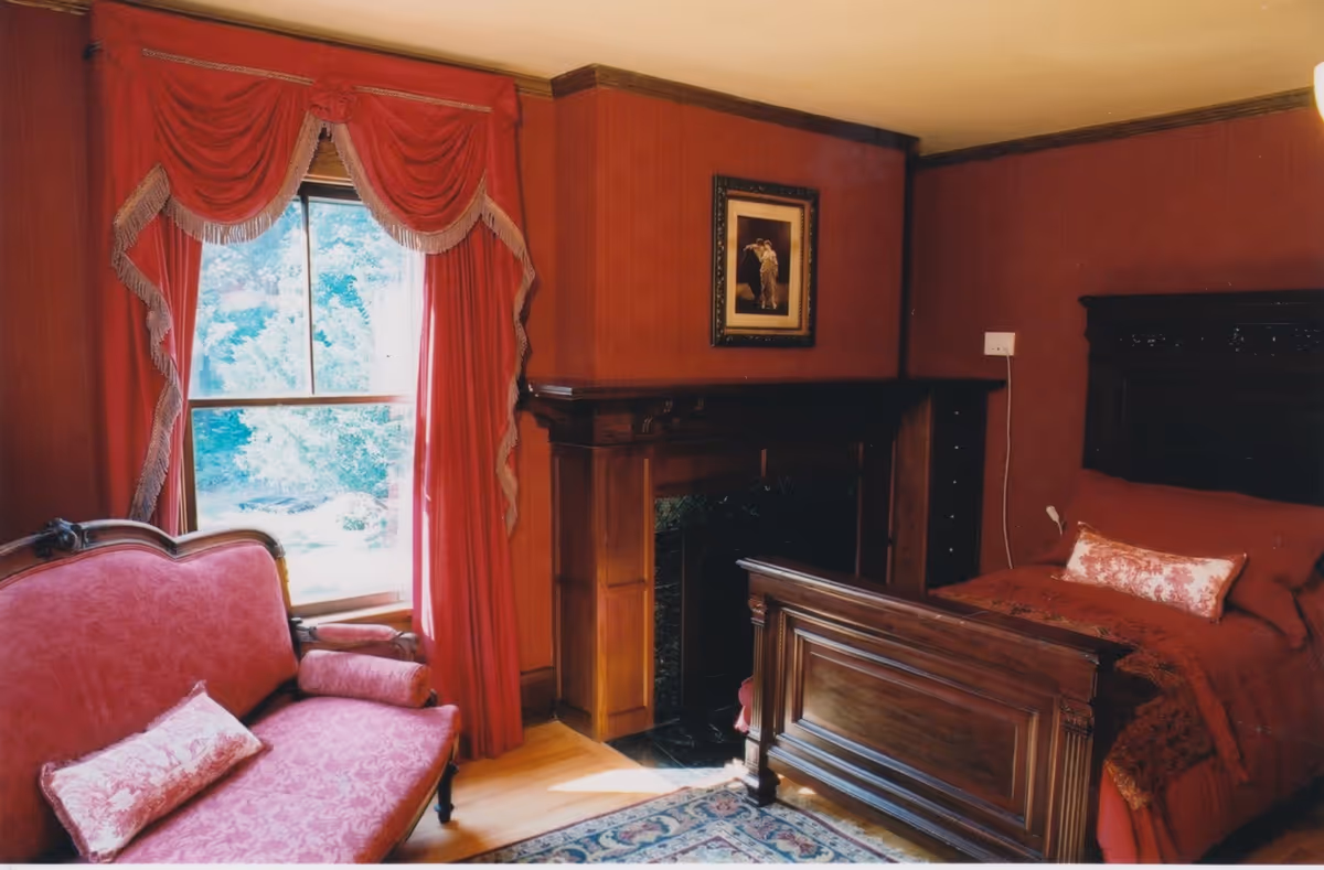 A cozy bedroom with dark wood furniture including a bed and a small sofa. The room features red walls and matching red curtains with decorative trim framing a window. There is a framed picture above a wooden fireplace mantel, and a patterned rug on the wooden floor.
