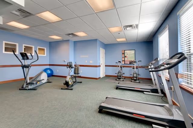 A fitness room with exercise equipment including two treadmills, two stationary bikes, and a blue exercise ball. The room has blue walls, carpeted floor, and large windows letting in natural light.