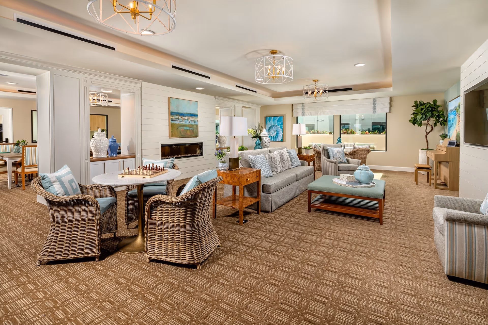 A bright and spacious living room area in Clearwater Newport Beach featuring comfortable wicker chairs with cushions around a small round table with a chessboard, a gray sofa with patterned pillows, a wooden coffee table with decorative items, armchairs, a piano, and large windows letting in natural light. The room is decorated with paintings, plants, and modern light fixtures on the ceiling.