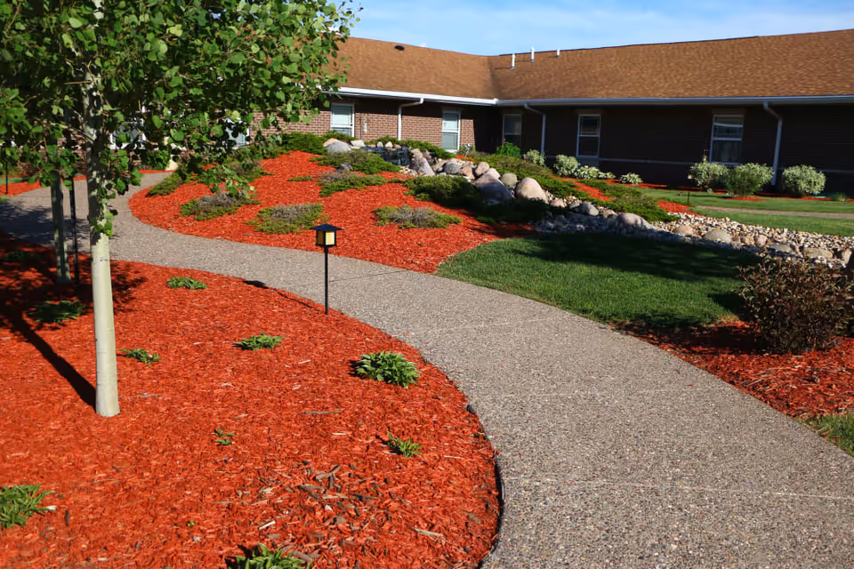 Curved concrete pathway winding through a landscaped garden area with red mulch, small green plants, rocks, and a tree, adjacent to a single-story brick building with a brown roof under a clear blue sky.