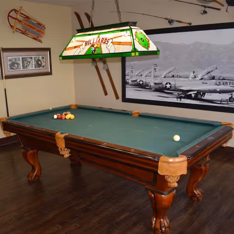 A billiards room featuring a green felt pool table with balls arranged on it, a stained glass hanging lamp above labeled 'Billiards', vintage wooden skis mounted on the wall, a framed black and white photo of U.S. Air Force planes, and a wooden floor.