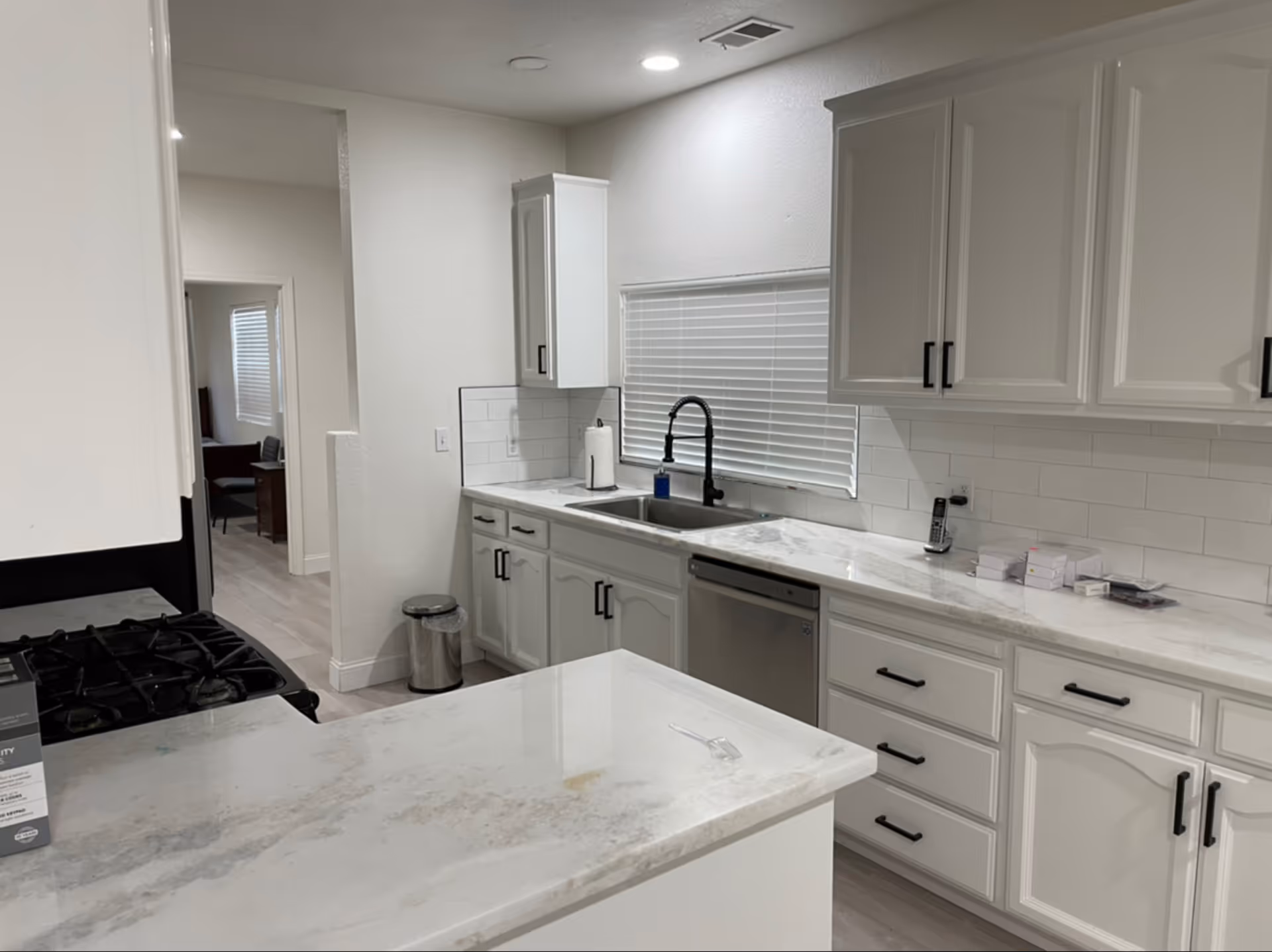 Modern kitchen with white cabinets, marble countertops, a stainless steel dishwasher, a black faucet over a double sink, a gas stove, and a window with white blinds. There is a small trash can on the floor and some items on the countertop including a phone and small boxes.