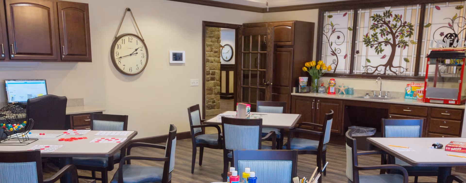 A cozy activity room in a senior living facility with several tables and chairs arranged for games and crafts. One table has bingo cards and red markers, another has board games like Yahtzee and pencils. The room features dark wood cabinets, a sink, a popcorn machine, and decorative stained glass windows with a tree design. A large wall clock and a computer workstation are also visible.
