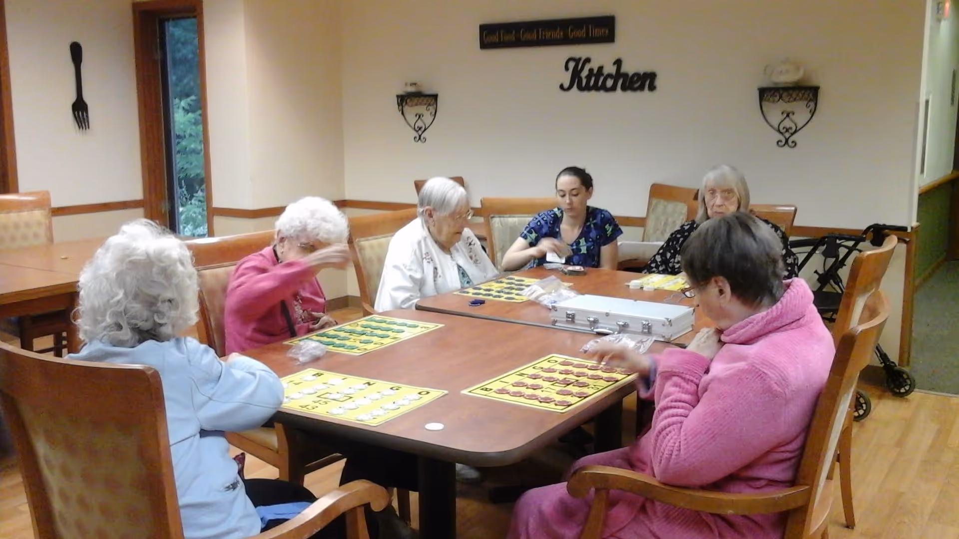 A group of elderly women and a caregiver sitting around a wooden table playing bingo in a room with beige walls. The wall has decorative signs including one that says 'Kitchen' and another that says 'Good Food - Good Friends - Good Times'. There is a window showing greenery outside and a walker is visible in the background.