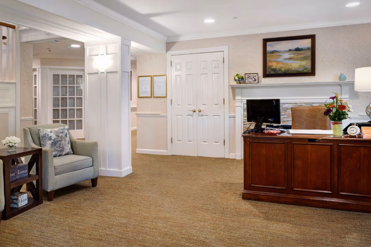 Bright reception lobby with a wooden front desk, computer, an upholstered armchair and side table, and framed artwork above a mantle.