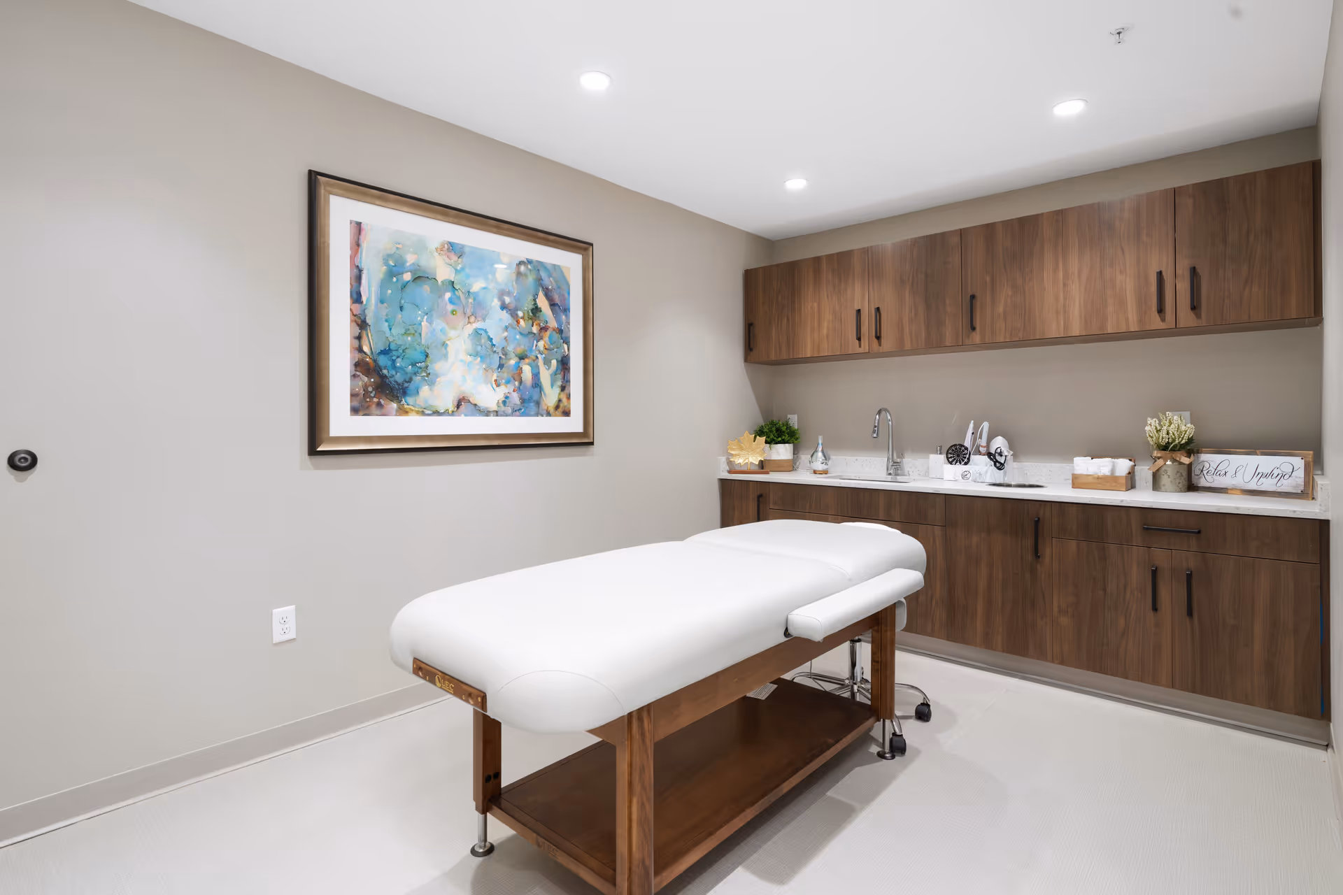 A clean therapy room with a white massage table, wooden cabinets, sink and a framed abstract painting on the wall.