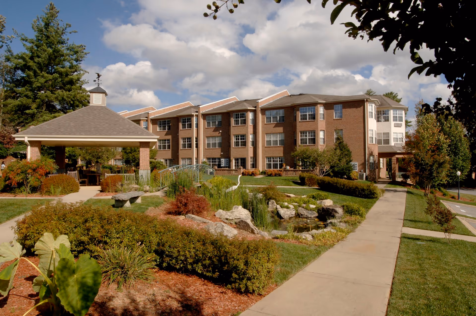 Outdoor view of Fletcher Park Inn Retirement showing a large brick building with multiple windows under a partly cloudy sky. In the foreground, there is a landscaped garden with bushes, rocks, a small pond, and a covered pavilion with seating. A paved walkway leads through the garden area toward the building.