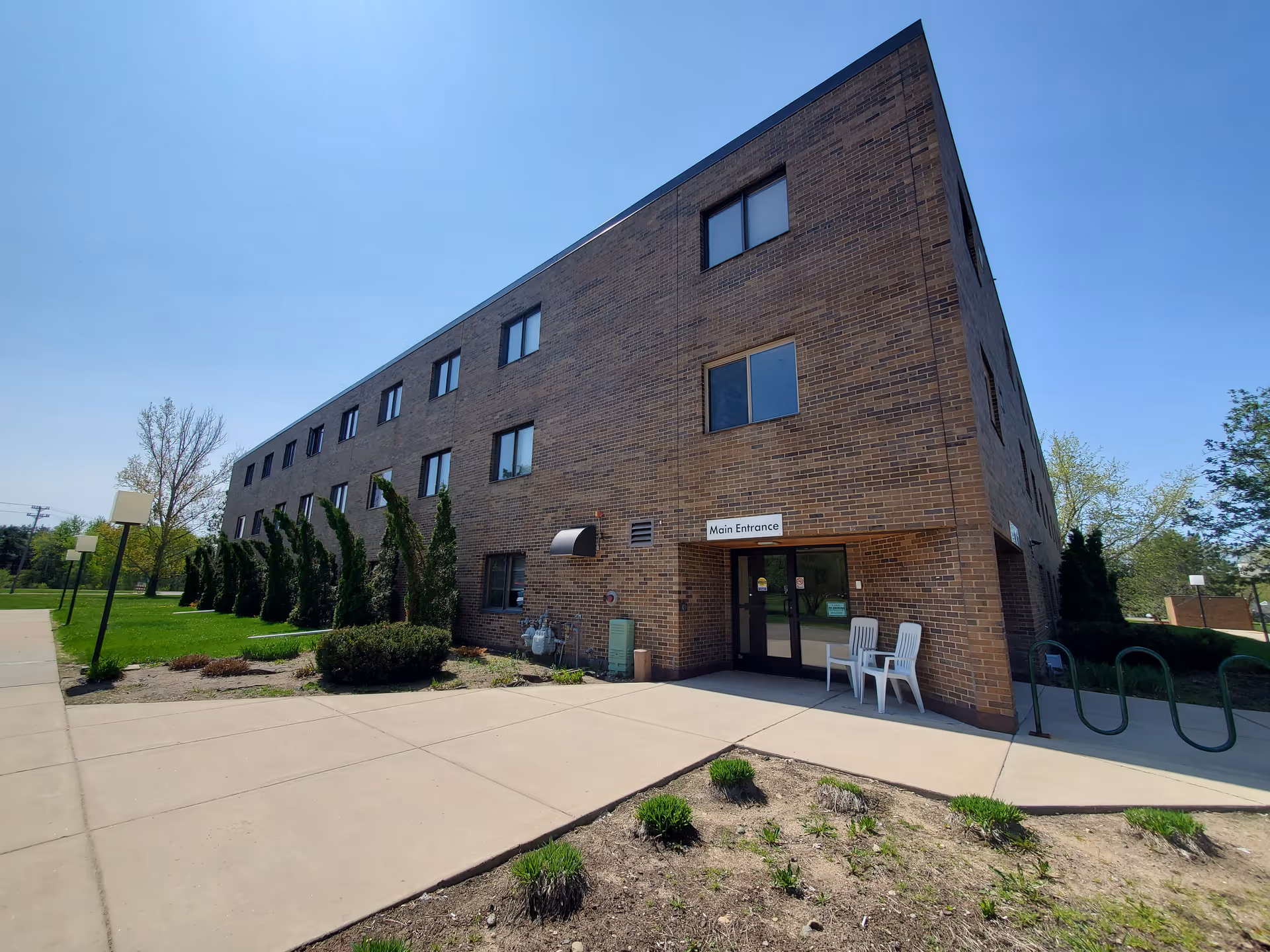 Brick senior living building with main entrance, sidewalk, and white chairs outside.