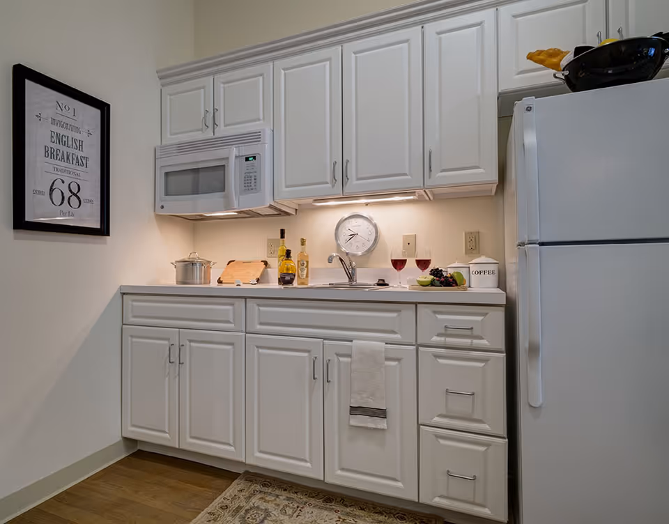 A compact kitchen area with white cabinets and drawers, a white microwave mounted above the countertop, a white refrigerator on the right, and a stainless steel sink. On the countertop, there are two glasses of red wine, a bowl of fruit, a coffee container, a cutting board, and two bottles of olive oil or vinegar. A clock is mounted on the wall above the sink, and a framed sign about English breakfast hangs on the left wall. The floor has a patterned rug partially visible.