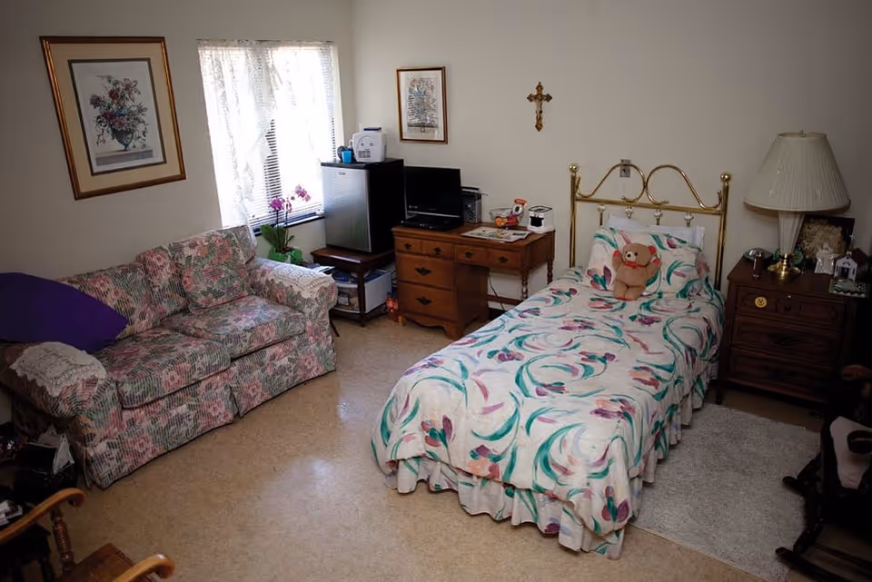 A small furnished bedroom with a single brass bed with a floral bedspread and teddy bear, a floral sofa, wooden dressers, lamp, and a window with curtains.
