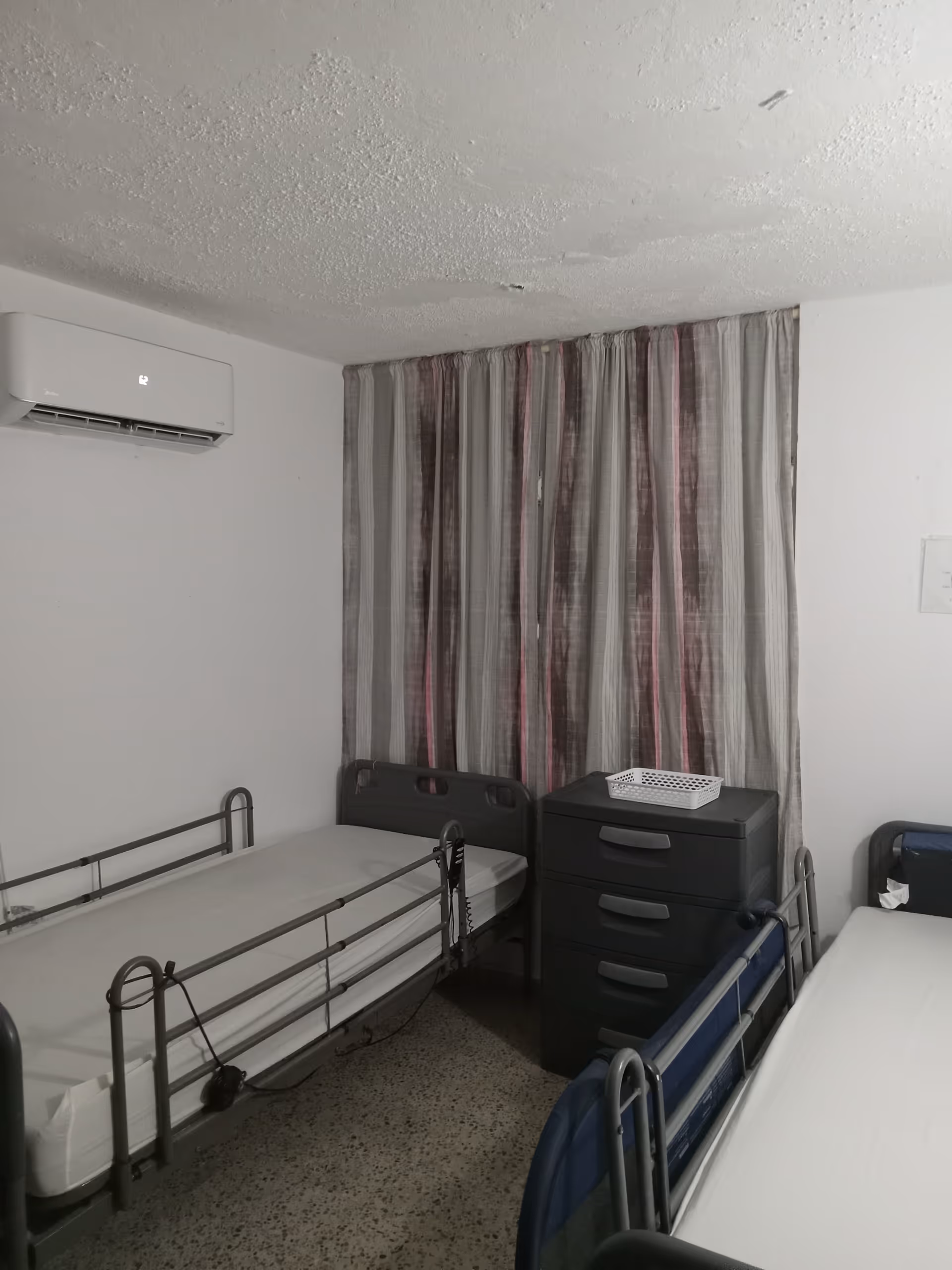 Shared bedroom with two hospital-style beds, a small dresser and striped curtains.