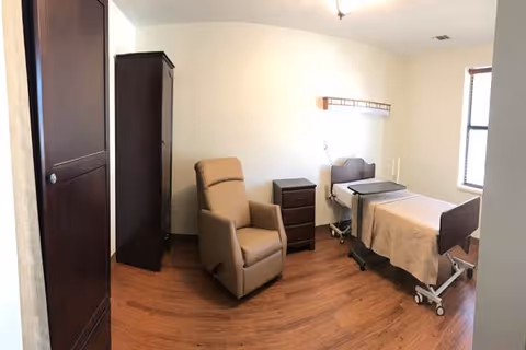 A simple, clean bedroom in a senior living facility featuring a hospital-style bed with beige bedding, a brown recliner chair, a small dark wooden nightstand, and a tall dark wooden wardrobe against a white wall. The room has wooden flooring and a window letting in natural light.