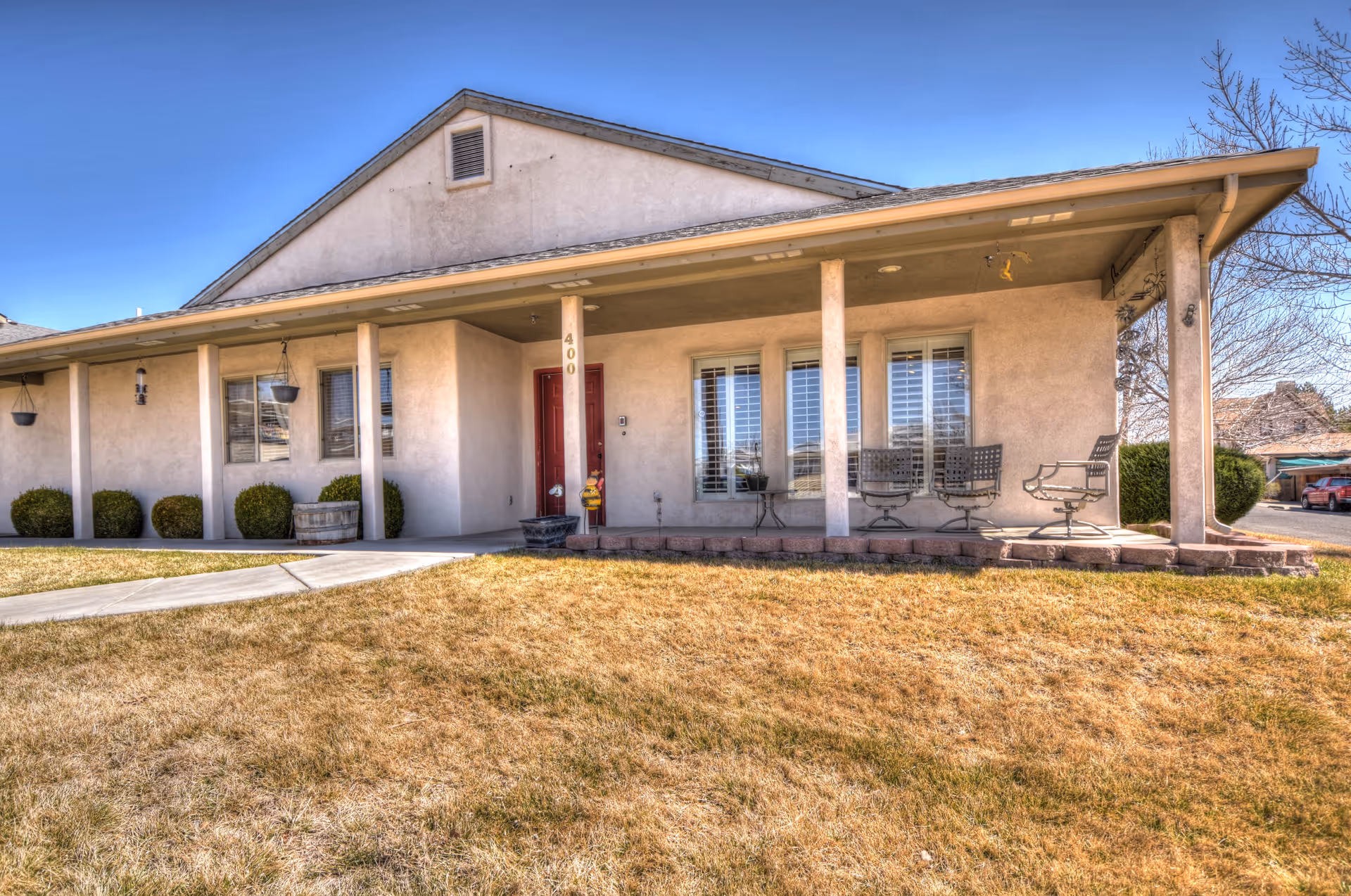 Exterior view of a single-story building with a covered porch supported by columns. The porch has several chairs and a small table. The building has a beige stucco finish, a red door, and several windows with white shutters. There is a dry grass lawn in front and a clear blue sky above.