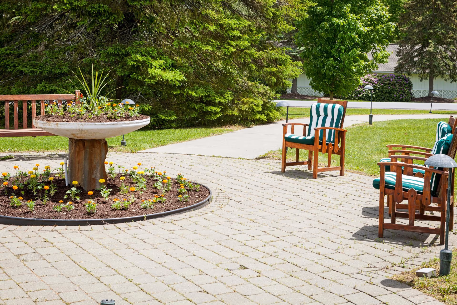 Sunlit outdoor patio with a circular flower planter, wooden chairs with green-and-white cushions, and surrounding lawn and trees.