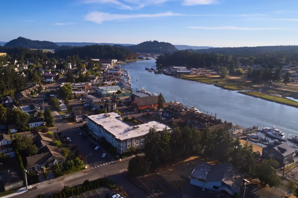 Aerial view of a small town along a river with boats docked at the waterfront, surrounded by trees and hills under a clear blue sky.