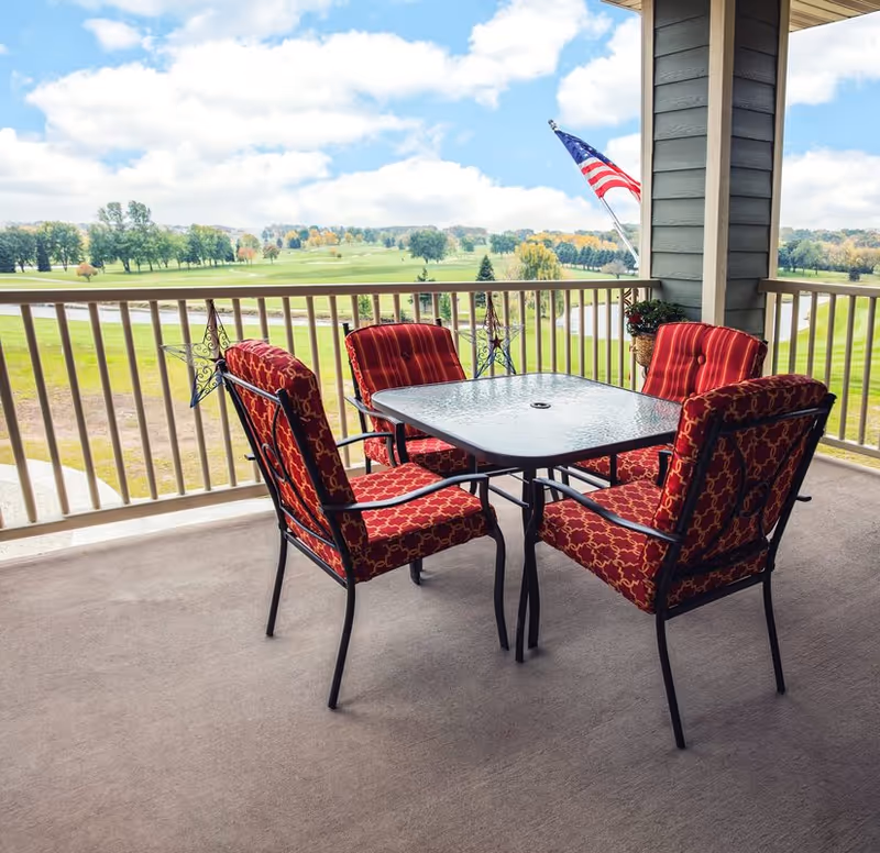 Balcony patio with a glass-top table and four red cushioned chairs overlooking a grassy golf course and an American flag.