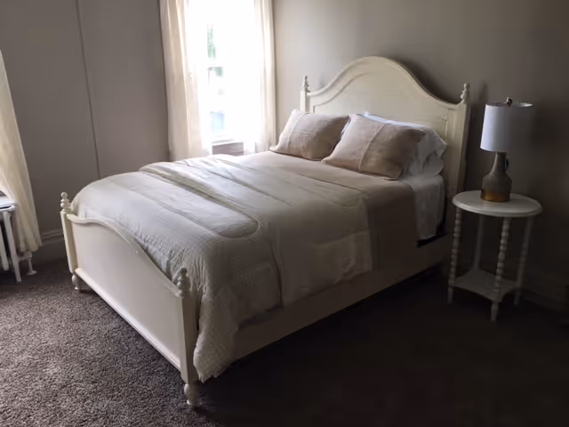 A cozy bedroom with a white wooden bed frame, neatly made with beige and white bedding and pillows. There is a small round white side table with a lamp on it next to the bed. Light filters in through a window with sheer curtains on the left side of the room.