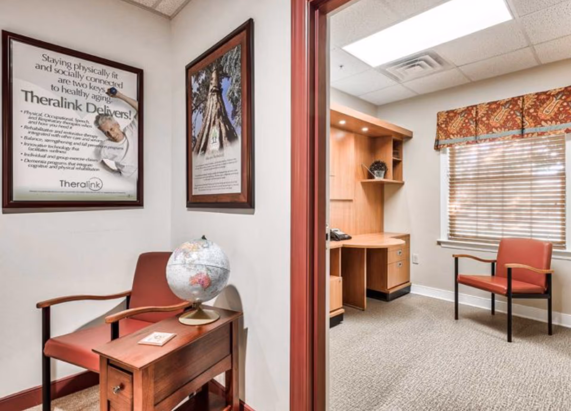 Interior view of a senior living facility office area with two red chairs, a small wooden table holding a globe, framed posters on the wall, a wooden desk with shelving, and a window with blinds and a patterned valance.