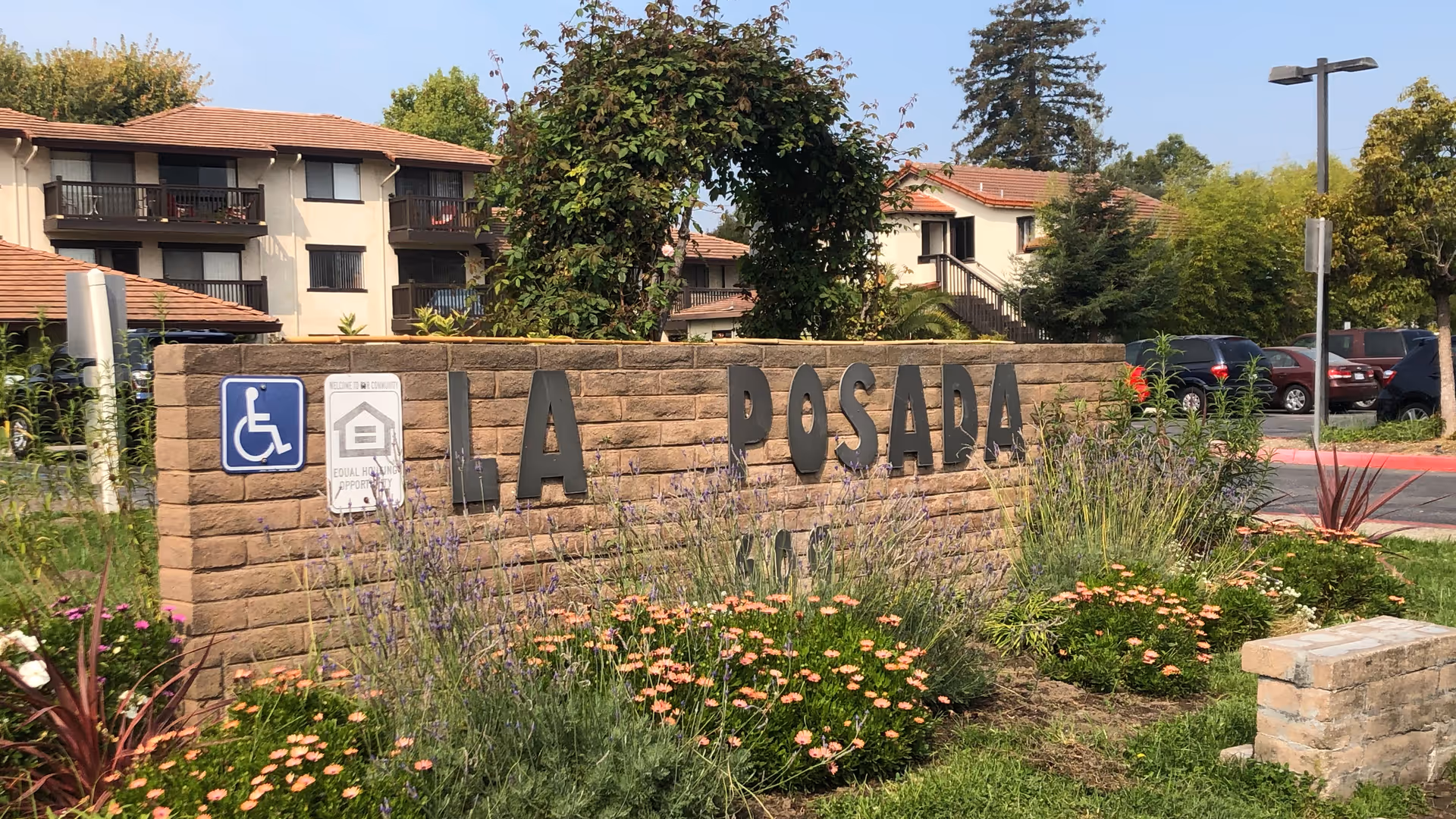 Outdoor view of the entrance sign for La Posada Retirement Community, a brick wall with large black letters spelling 'LA POSADA' surrounded by landscaped flowers and greenery, with apartment buildings and parked cars in the background under a clear blue sky.