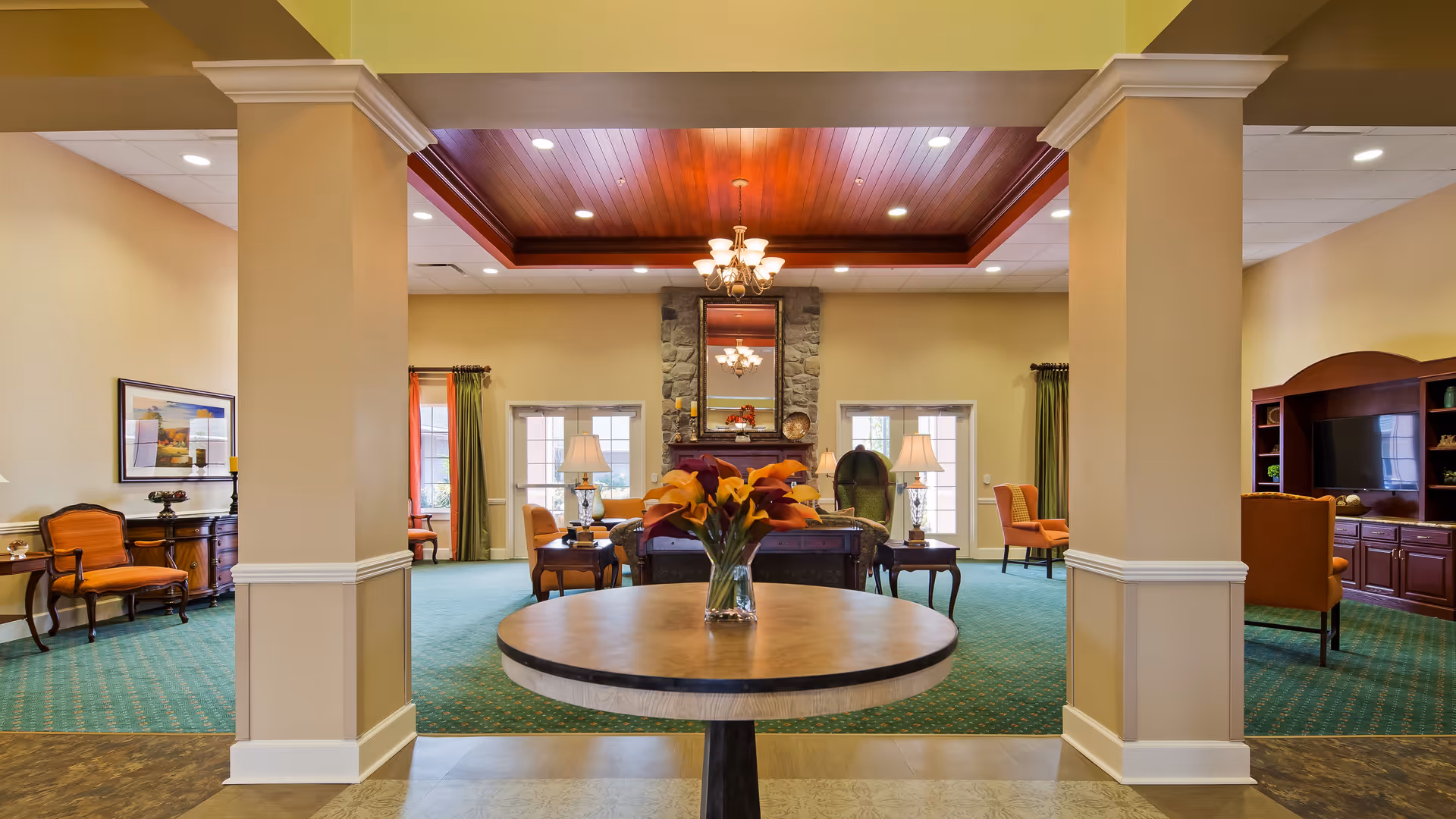 Spacious senior living facility common area with green carpet, beige walls, and a wooden ceiling accent. The room features a round table with a vase of flowers in the foreground, multiple seating areas with chairs and sofas, lamps, a stone fireplace with a mirror above it, and a large wooden entertainment center with a TV.