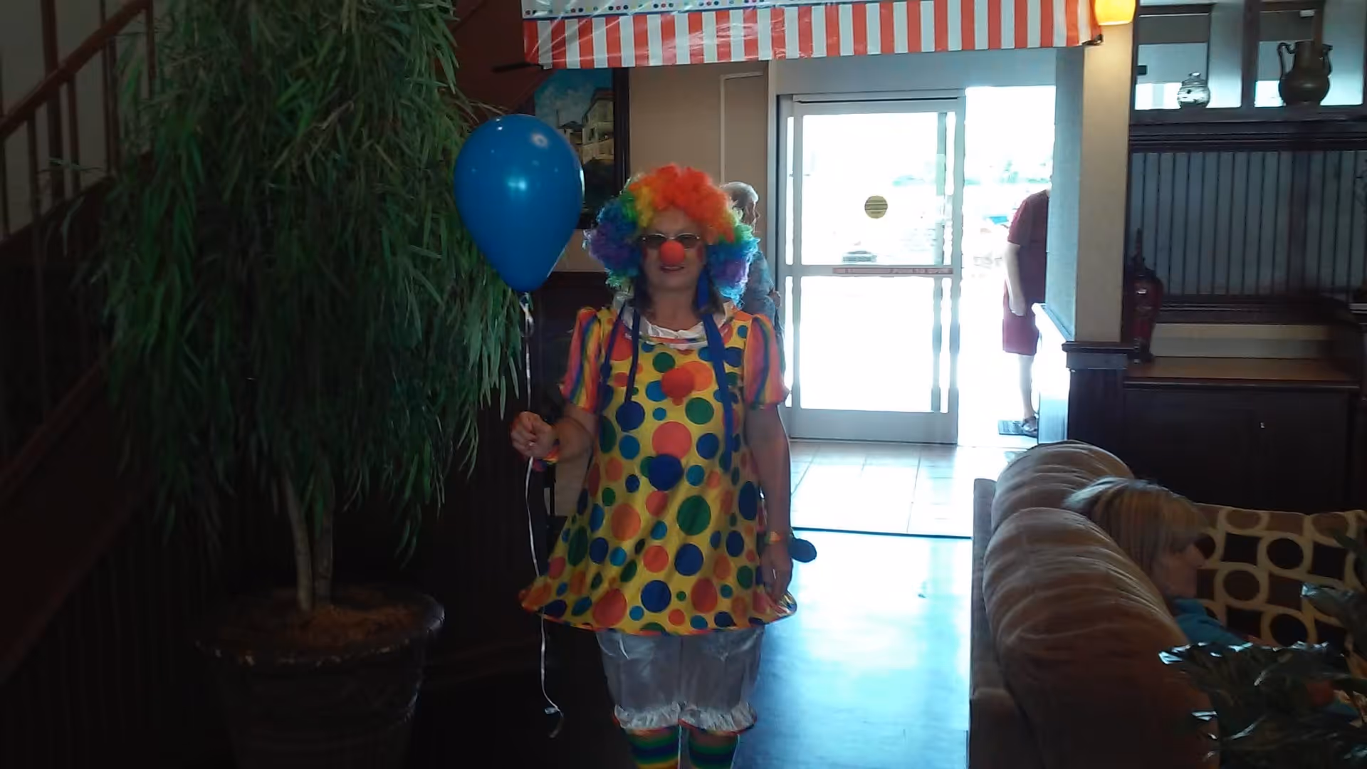 A person dressed as a colorful clown holding a blue balloon standing inside a senior living facility lobby area. There is a large potted plant to the left, a staircase railing behind, and a seating area with a person sitting on a couch to the right. The entrance door with glass panels is visible in the background.