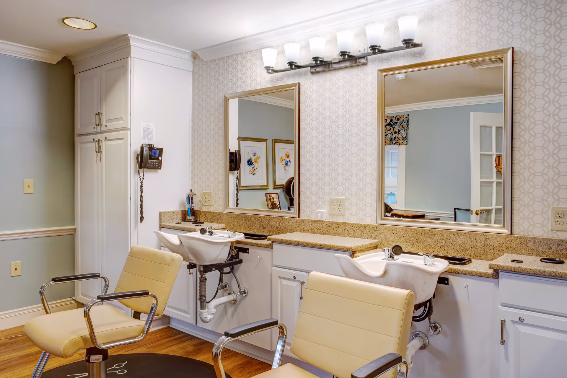 Interior view of a hair salon area with two beige salon chairs in front of two white sinks with mirrors above them. The room has light-colored walls with patterned wallpaper behind the sinks, wooden flooring, and white cabinetry. There is a wall-mounted phone on the left side and framed artwork reflected in the mirrors.