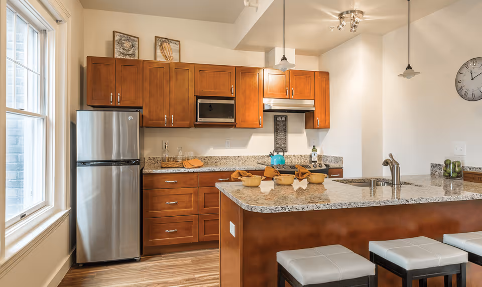Modern kitchen with wooden cabinets, granite countertops, a stainless steel refrigerator, microwave, stove, and a kitchen island with three cushioned stools. The kitchen is well-lit with natural light from a large window and ceiling lights.