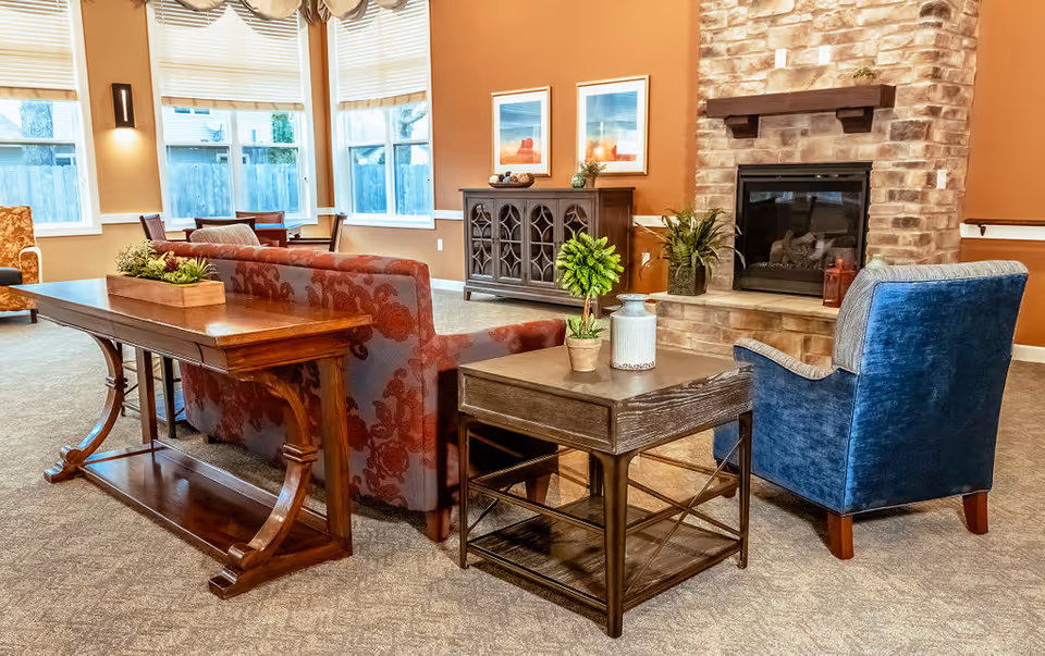 A cozy living room area with a red patterned sofa, a blue armchair, and wooden tables with decorative plants. The room features a stone fireplace with a mantel, two framed pictures on the wall, large windows with blinds, and warm lighting.