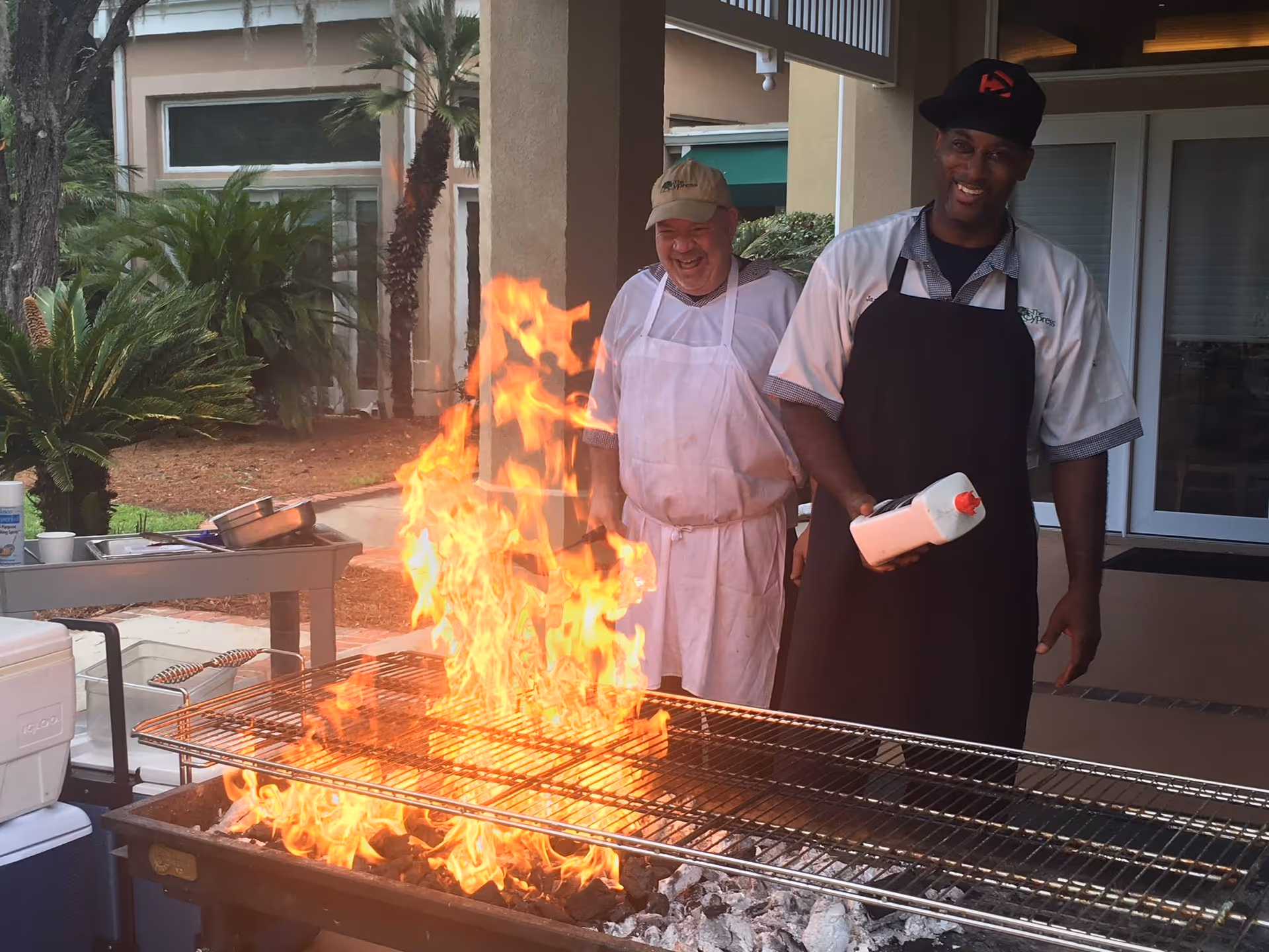 Two men in aprons stand behind a large outdoor grill with tall flames during a cookout at the facility.